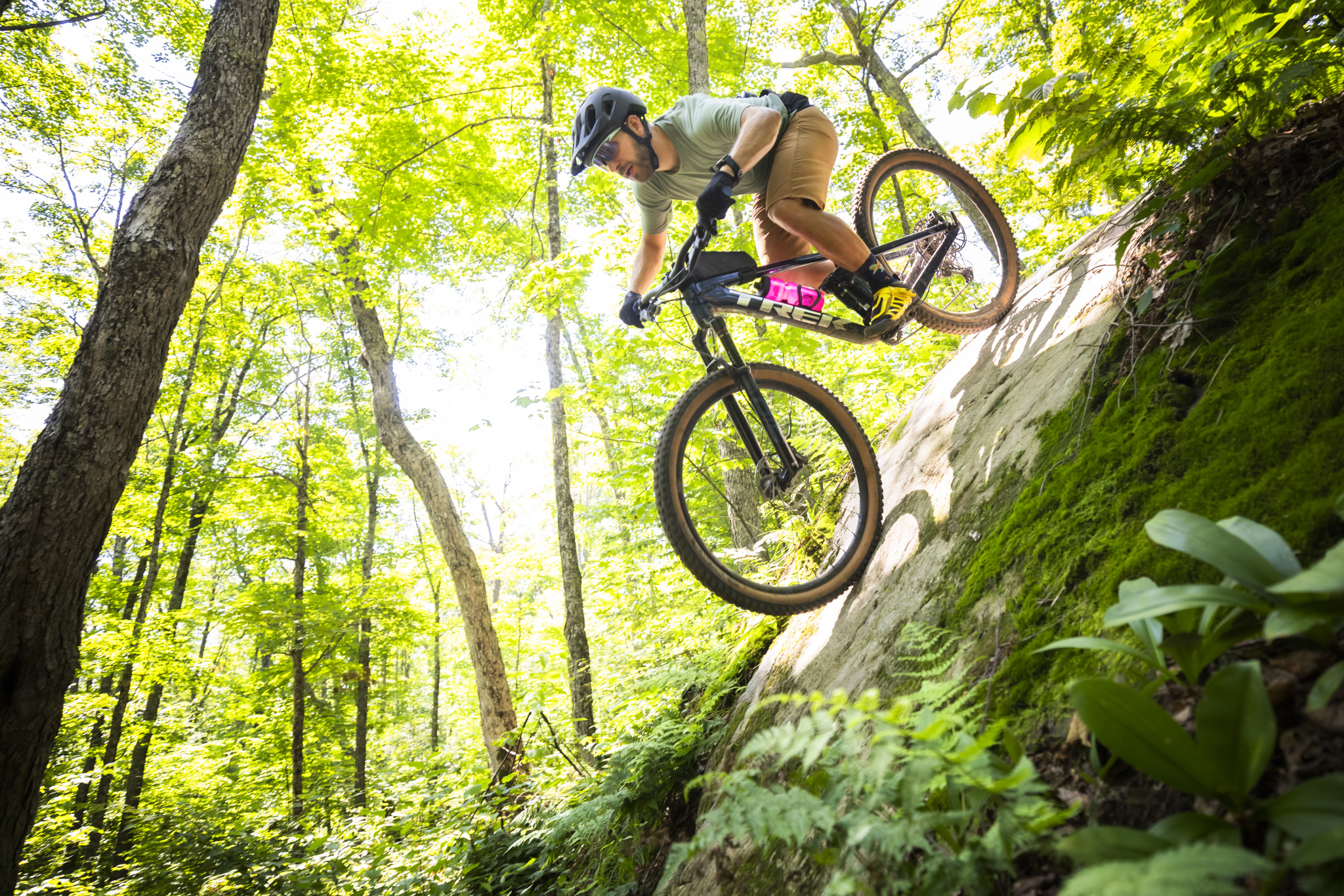 A person in a helmet and casual attire rides a mountain bike down a rocky incline in a lush, green forest. Sunlight filters through the trees, creating a vibrant backdrop of leaves and foliage. The rider appears focused and in motion, showcasing an adventurous moment in a natural outdoor setting.