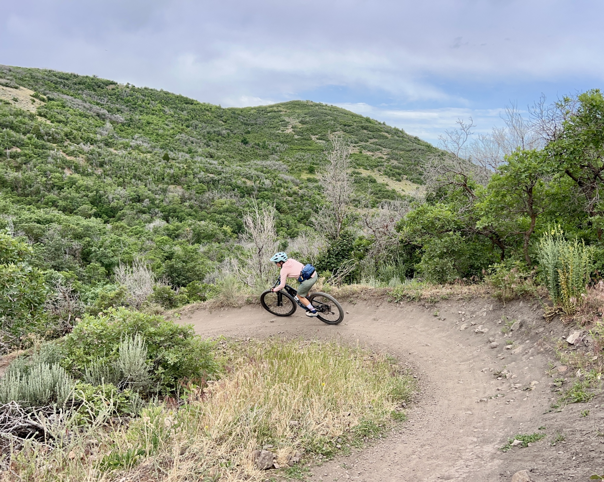 A person riding a mountain bike around a curved dirt trail on a hillside, surrounded by lush green vegetation and gentle slopes under a partly cloudy sky.