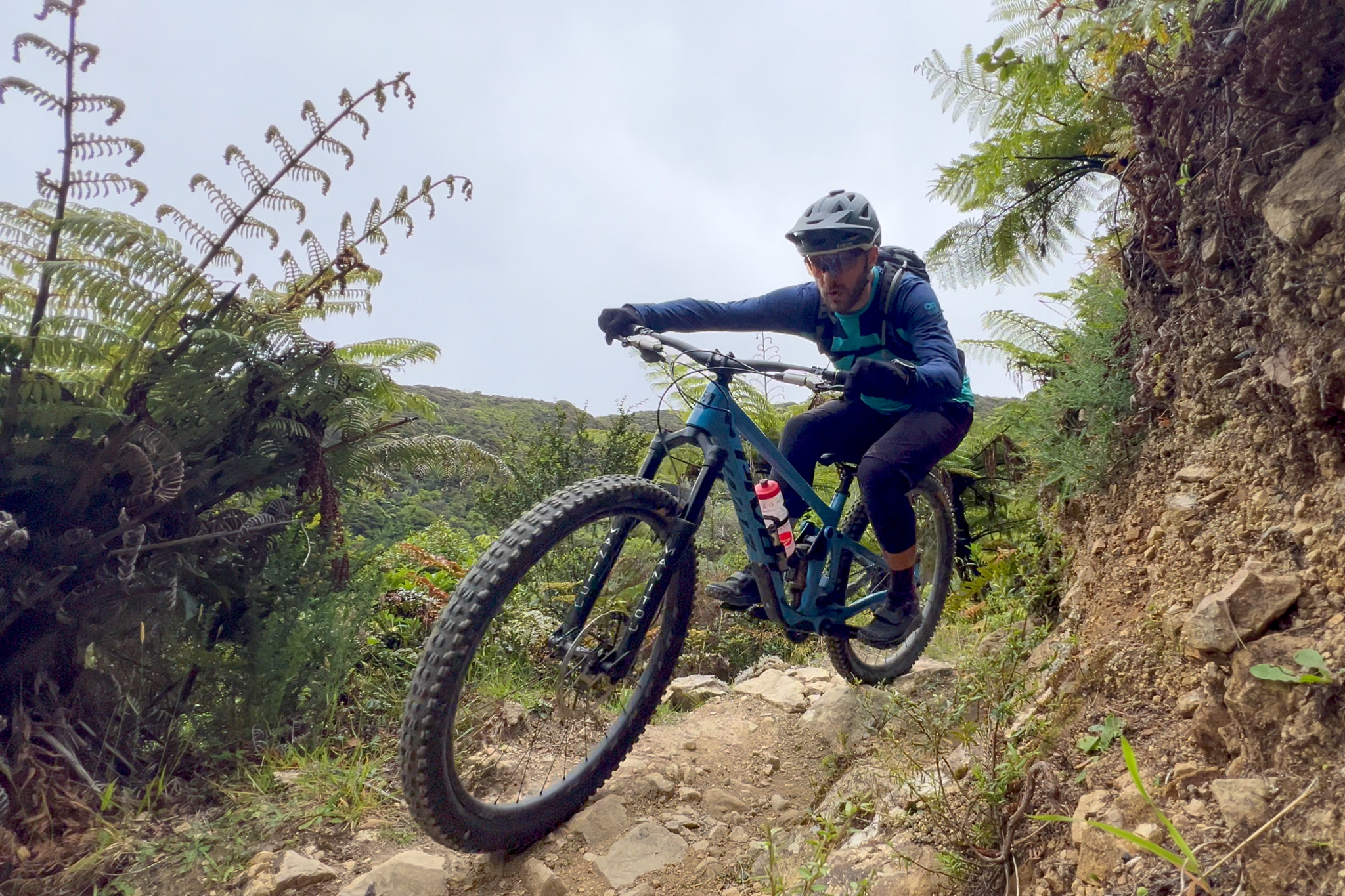 A mountain biker skillfully maneuvers down a rocky and uneven trail surrounded by lush ferns and greenery. The rider is wearing a helmet and sports gear, focusing intently as they navigate the terrain.