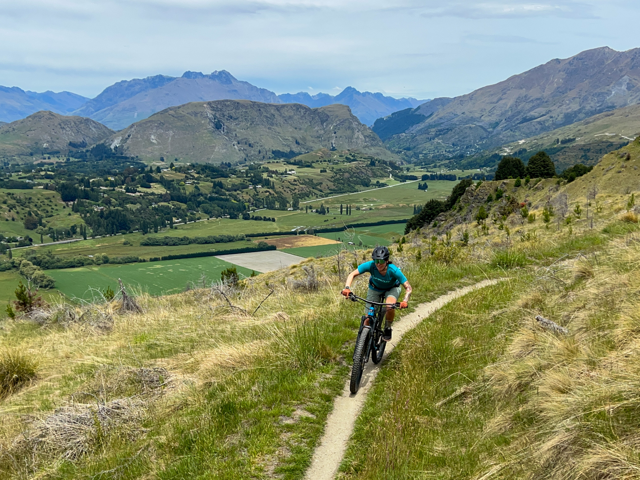 A mountain biker navigating a dirt trail on a hillside with lush green grass, surrounded by mountains and valleys in the background, under a partly cloudy sky.