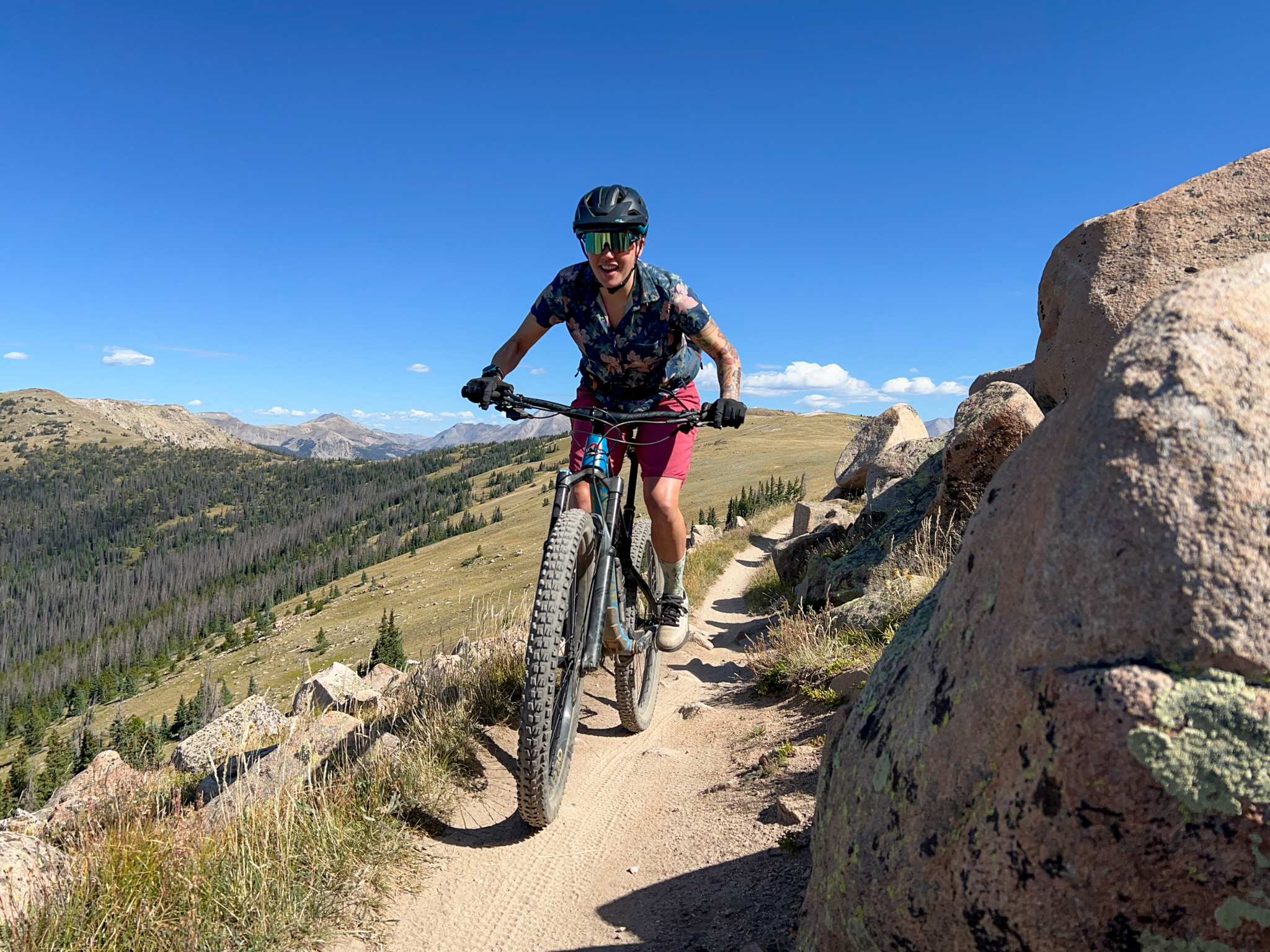 A mountain biker riding along a narrow dirt trail surrounded by rocky terrain and greenery. The scene features a clear blue sky and mountains in the background, highlighting an adventurous outdoor setting. The cyclist is wearing a helmet and vibrant athletic clothing.