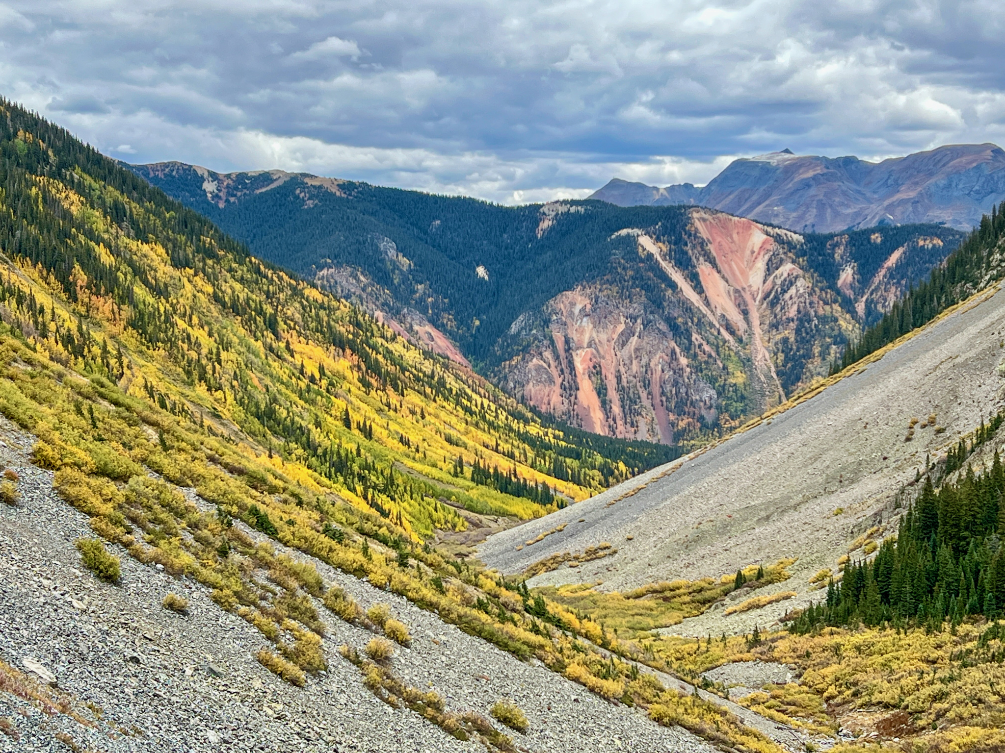 A scenic view of a mountainous landscape featuring rolling hills covered in vibrant autumn foliage, with patches of yellow and green trees. The foreground shows rocky terrain, while the background displays steep mountains and a cloudy sky, lending a dramatic atmosphere to the scene.