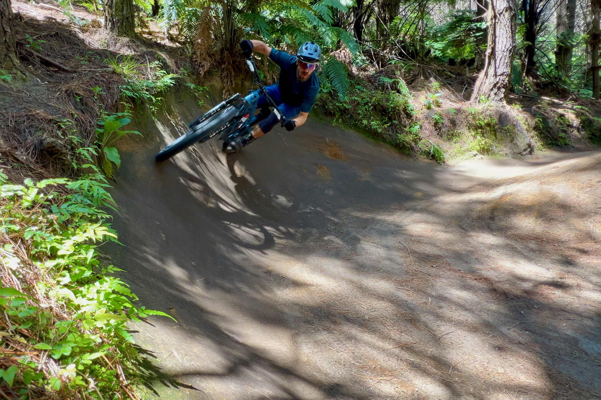 A mountain biker navigating a dirt trail, leaning into a curve surrounded by lush greenery and trees in a forest setting.
