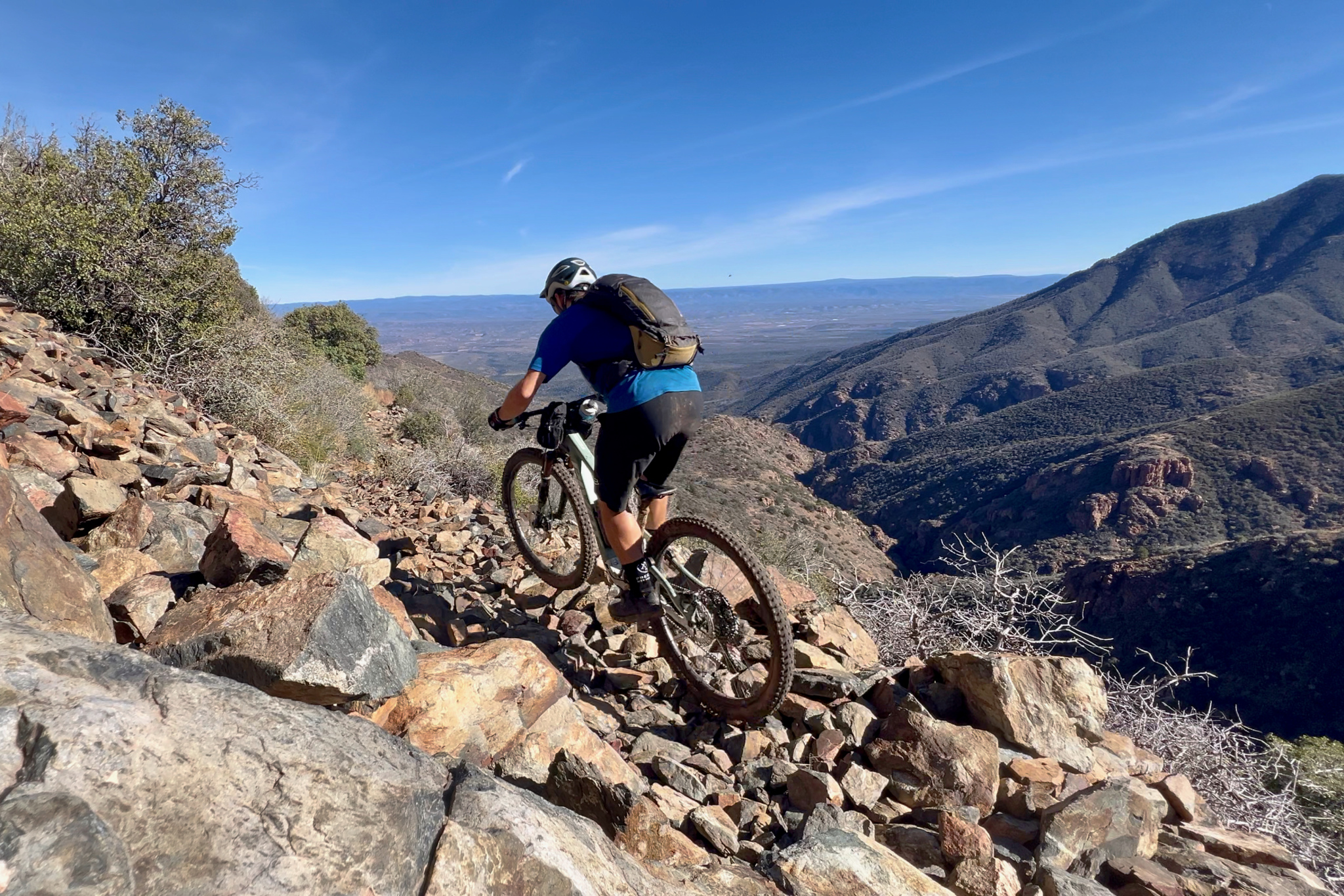 A mountain biker navigating a rocky trail on a hillside, with a panoramic view of mountains and valleys in the background under a blue sky.