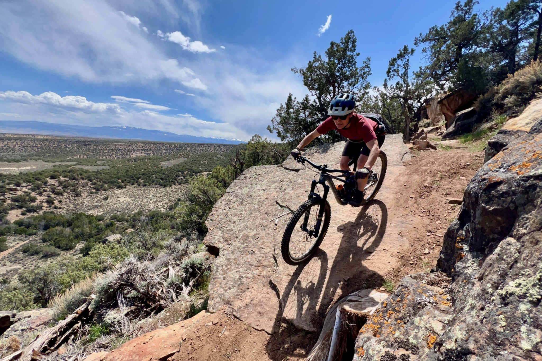 A mountain biker riding on a rocky trail with a scenic view of rolling hills and a blue sky filled with clouds. The terrain is rugged, with shrubs and trees lining the path. The biker is wearing a helmet and appears focused on navigating the trail.