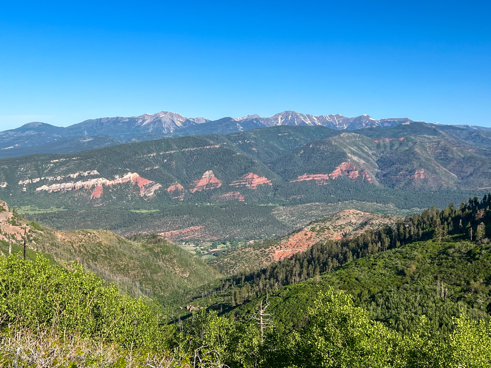 A panoramic view of a mountainous landscape featuring layered red rock formations, lush greenery, and a clear blue sky. The scene captures a mix of slopes and valleys, with distant snow-capped peaks appearing in the background. Haflin Creek mountain bike trail.