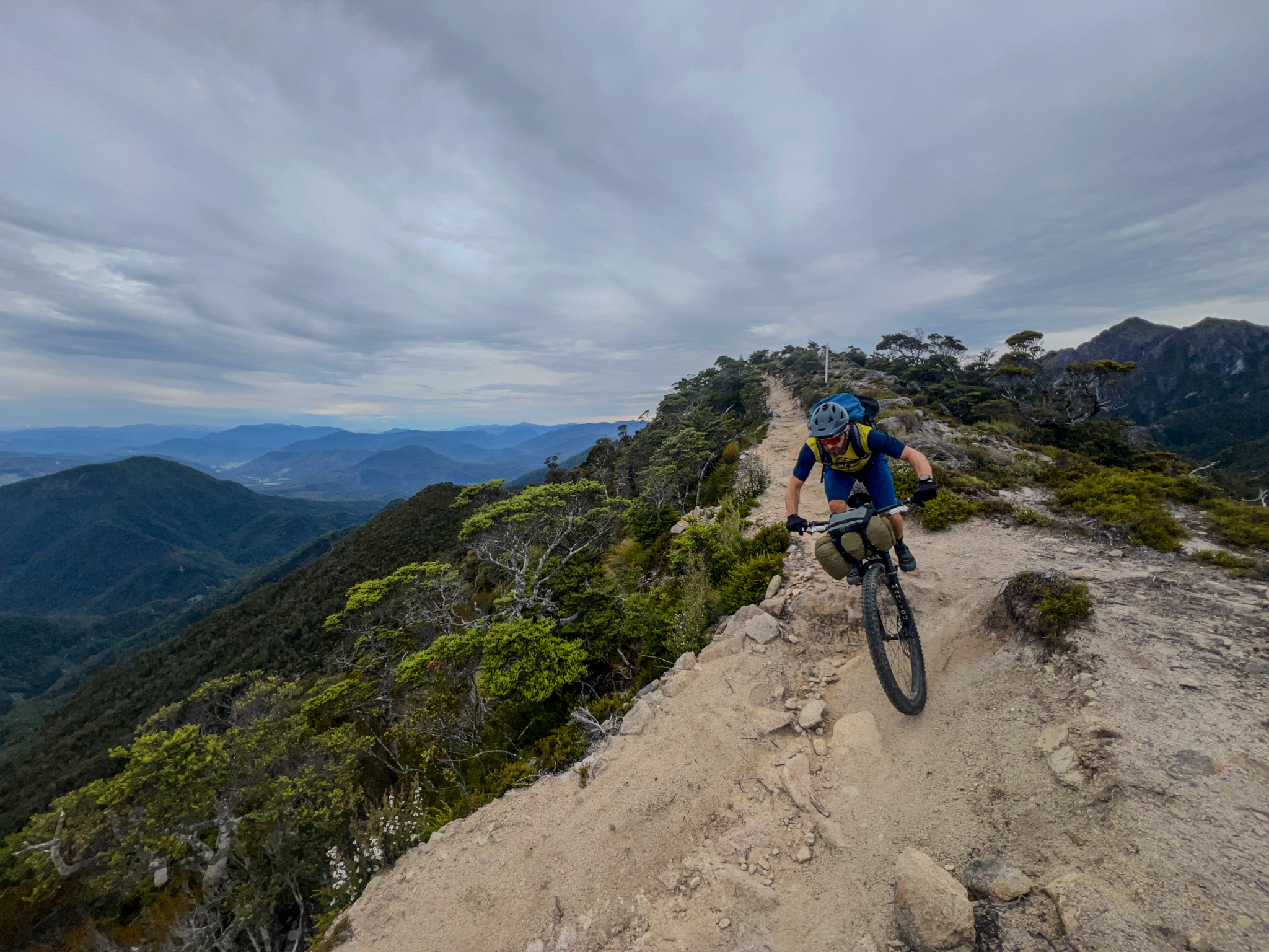 A mountain biker navigates a narrow, rugged trail along a ridge, surrounded by lush greenery and distant mountains under a cloudy sky. The cyclist wears a helmet and has a backpack, focused on the path ahead.