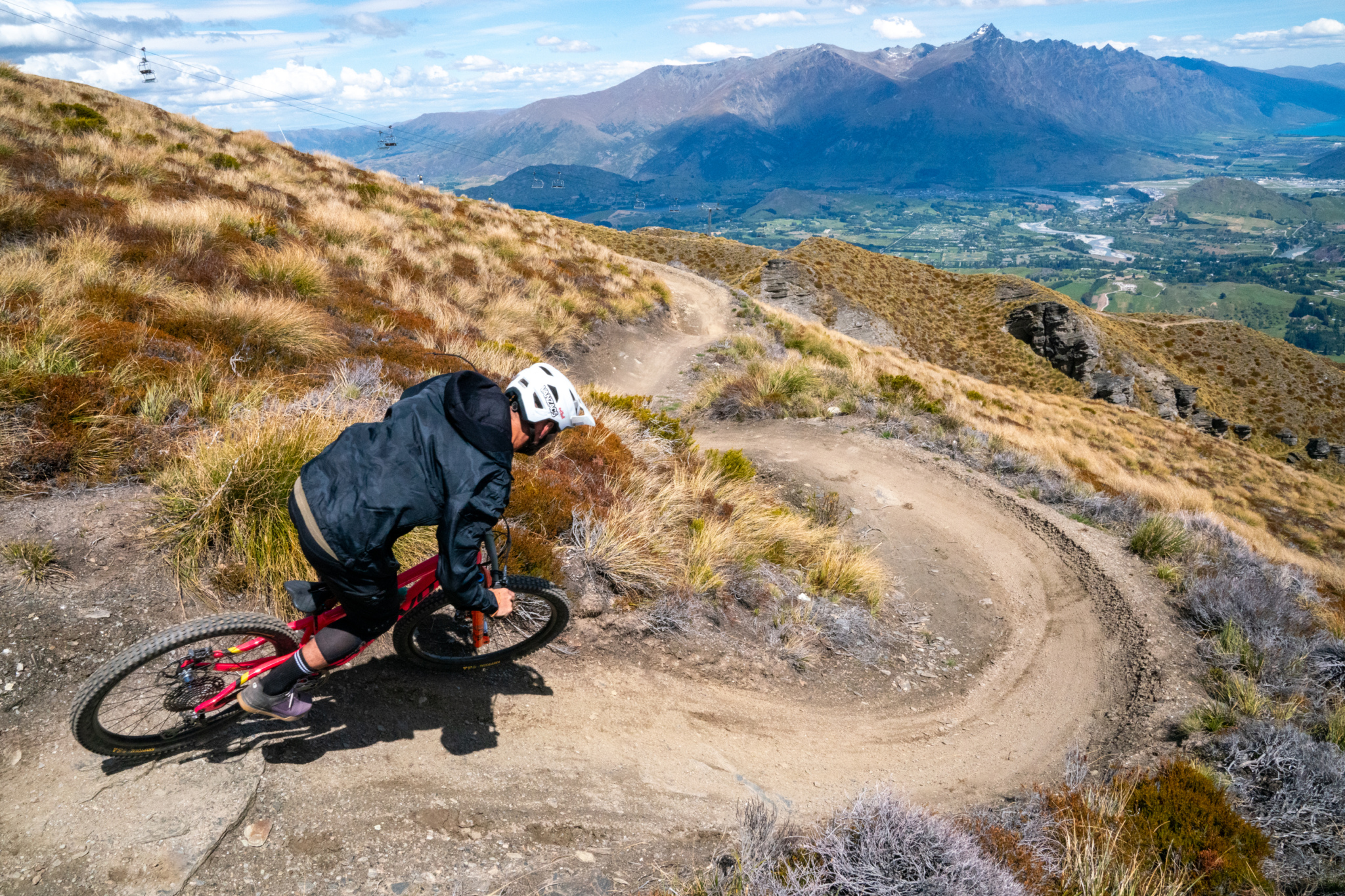 Mountain biker navigating a winding dirt trail on a hillside, surrounded by grass and shrubs, with a picturesque mountain landscape in the background under a blue sky.