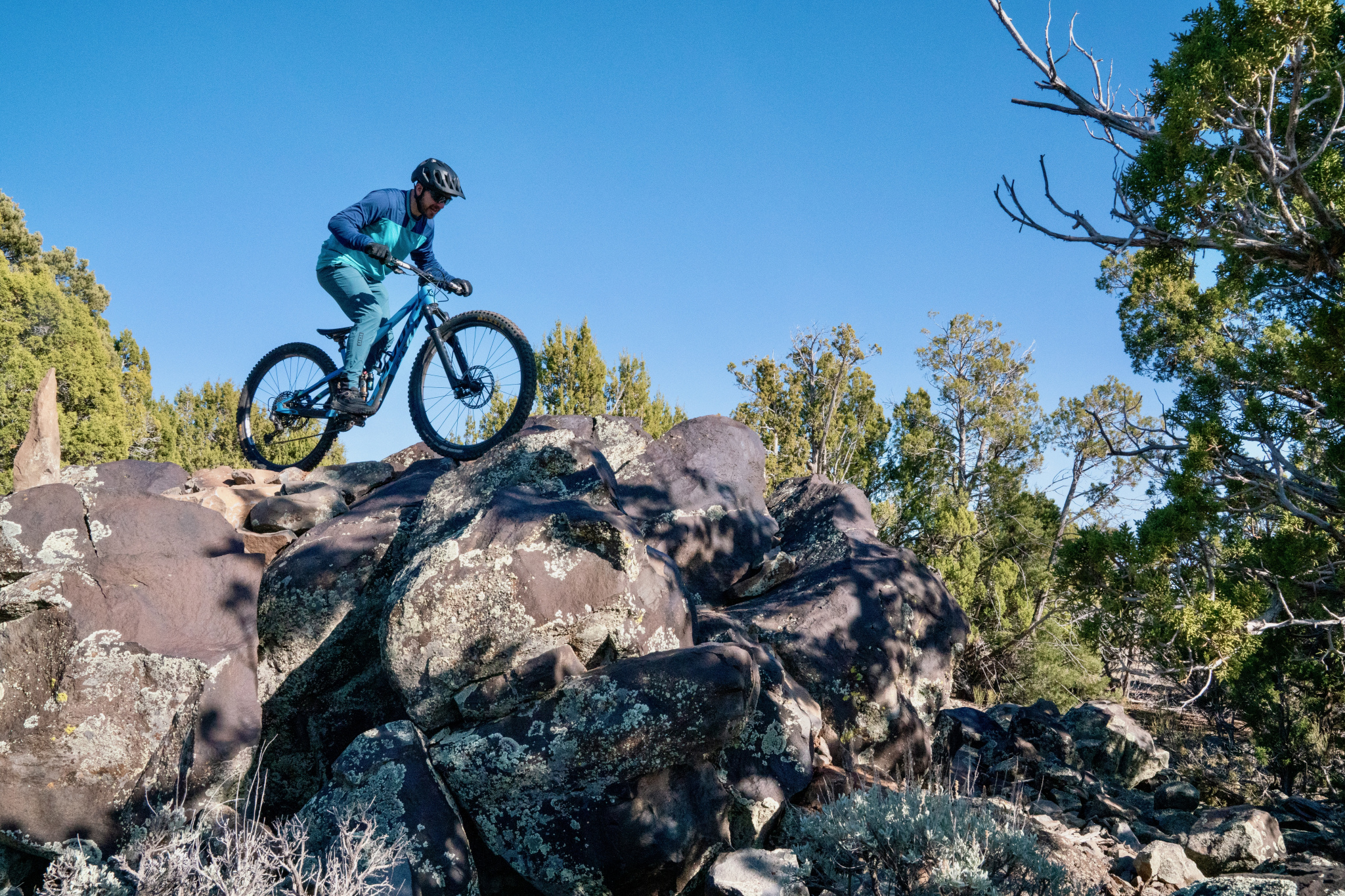 A mountain biker wearing a blue and teal outfit navigates over large rocks in a natural outdoor setting with trees and clear blue sky in the background.