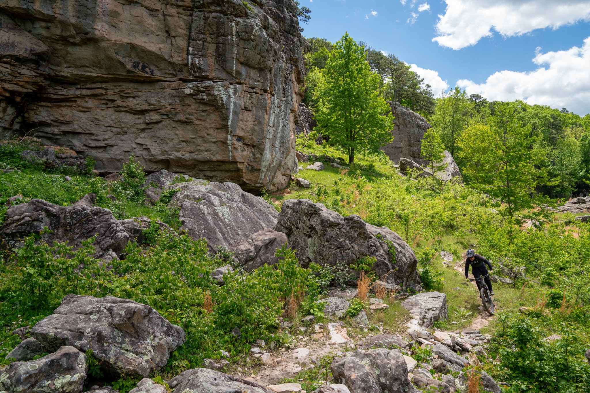 A mountain biker navigating a rocky trail in a lush green landscape, surrounded by large boulders and trees under a partly cloudy sky.