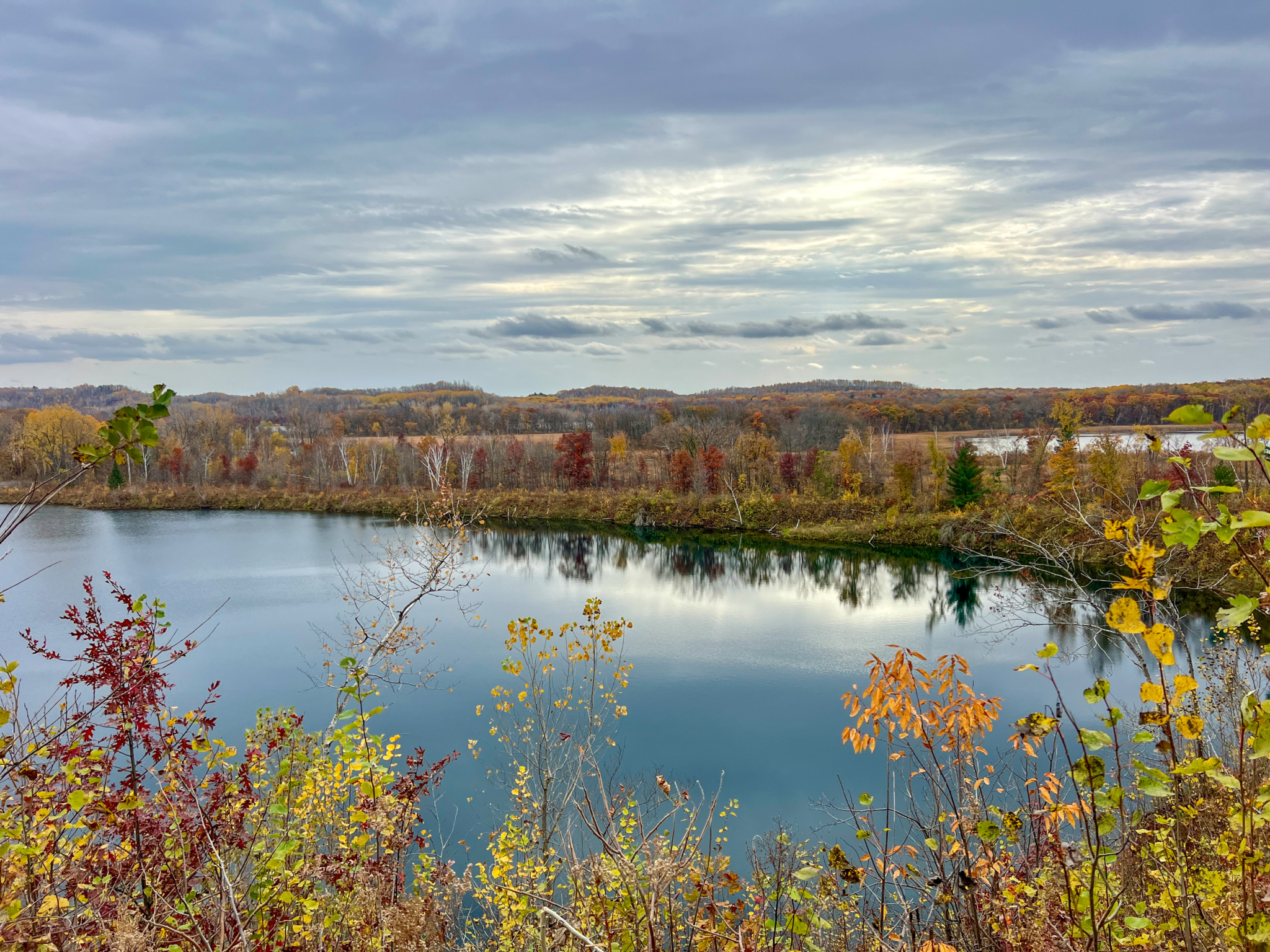 A serene view of a calm river surrounded by autumn foliage, featuring vibrant colors in the trees. The sky is overcast with soft gray clouds, reflecting the peaceful mood of the scene. The landscape includes rolling hills in the background and lush greenery in the foreground.