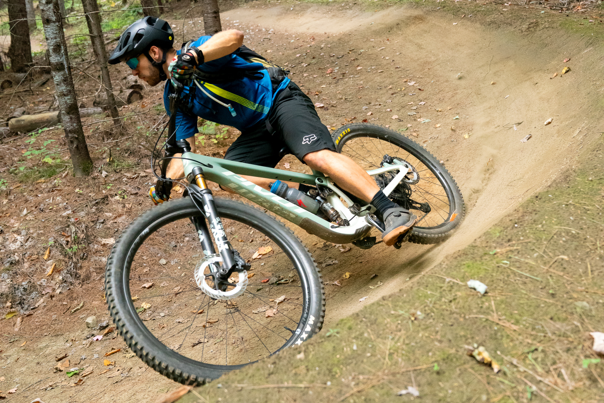 A mountain biker leans into a turn on a dirt trail surrounded by trees, showcasing an action-packed moment of riding. The cyclist is wearing a helmet, a blue shirt, and black shorts, with a water bottle attached to the bike frame. Dust is kicked up from the trail, adding to the dynamic feel of the scene.