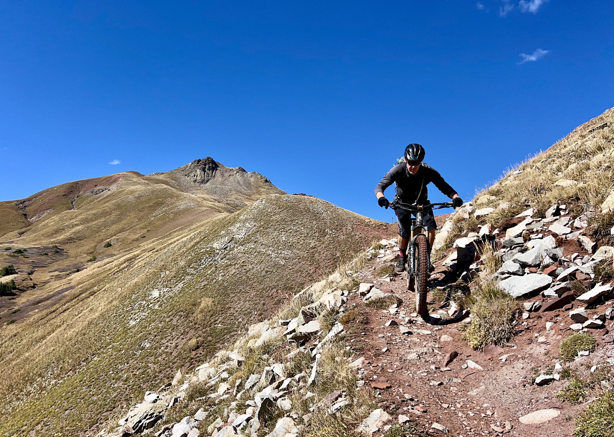 A mountain biker descends a rocky singletrack traversing a steep, grassy alpine ridge under a deep blue sky, with a jagged peak rising behind.