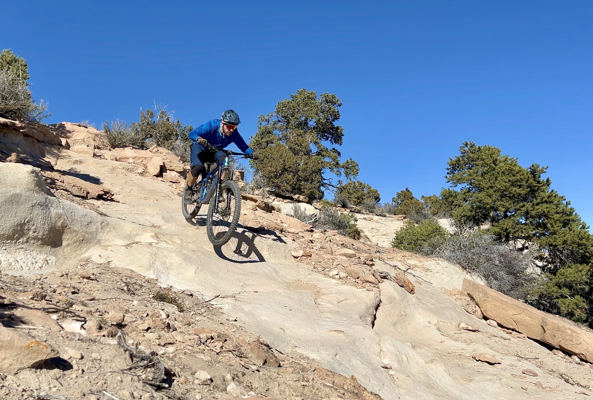A rider in a blue jacket drops down a broad slab of pale sandstone slickrock, scattered junipers and clear blue sky behind.