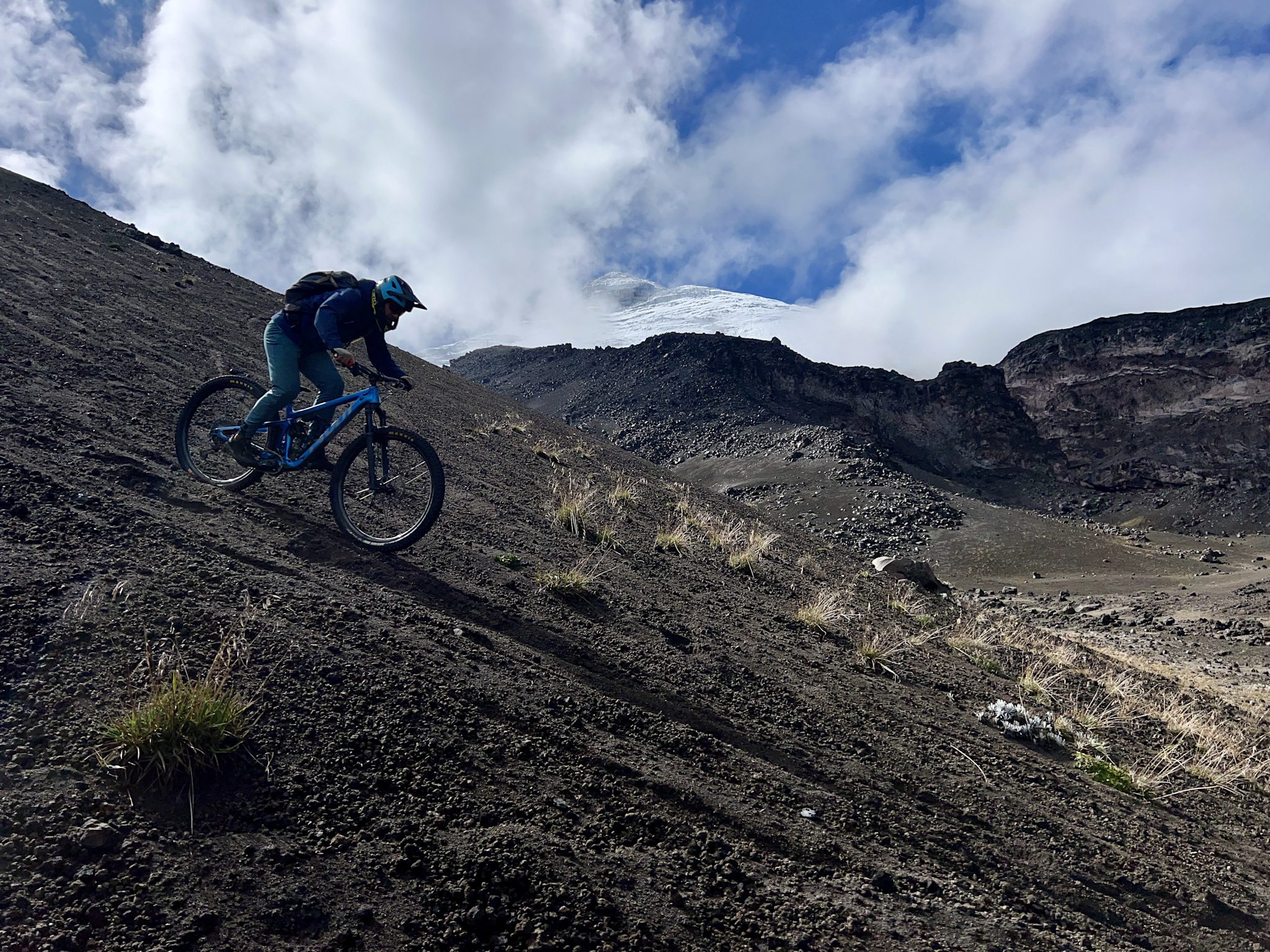 A rider descends a steep slope of black volcanic gravel, with the glaciated summit of a snow-capped volcano emerging from clouds behind.