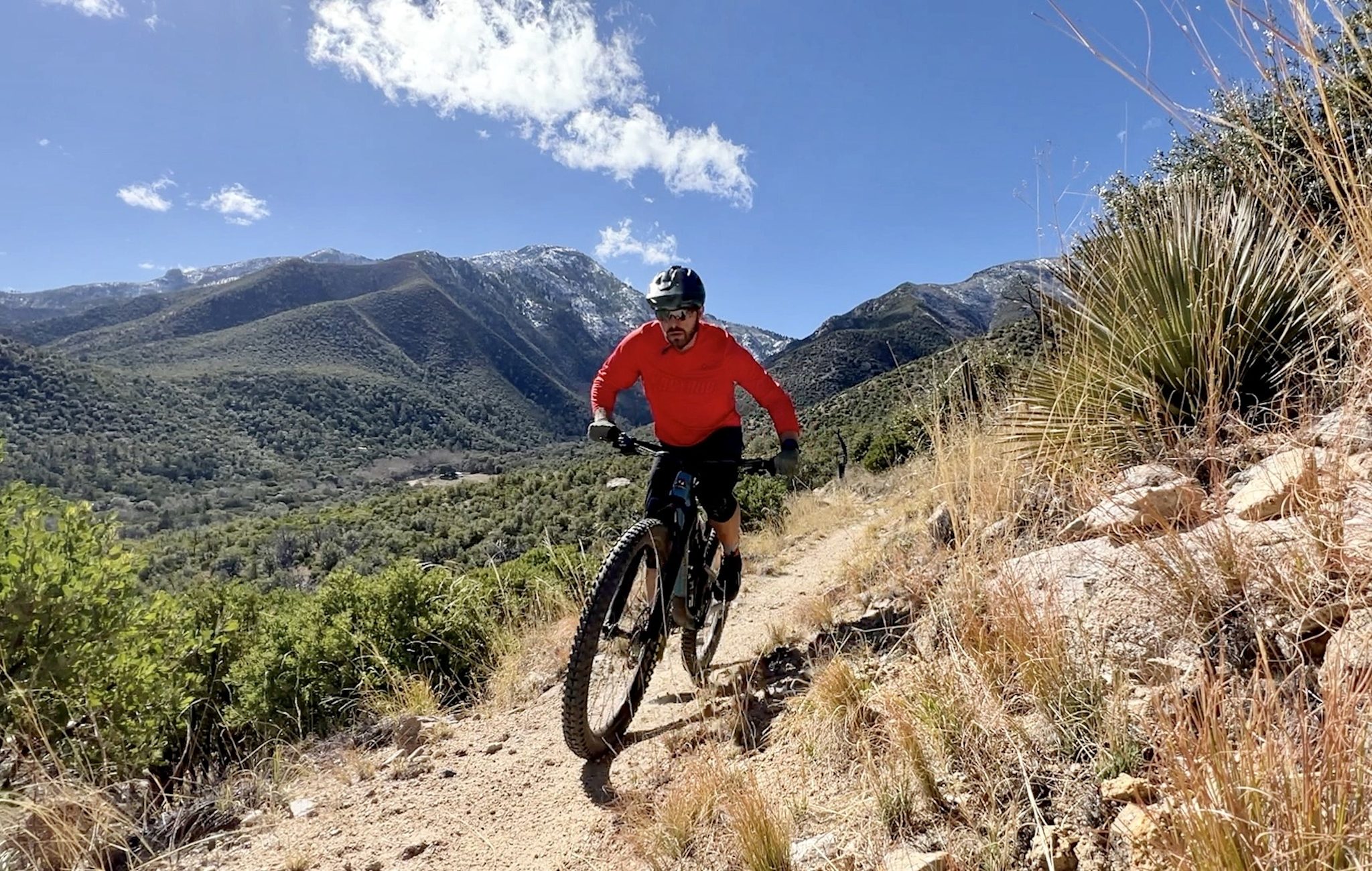 A mountain biker in a red jersey rides a rocky trail through high desert scrub, with a snow-dusted sky-island mountain range rising behind.
