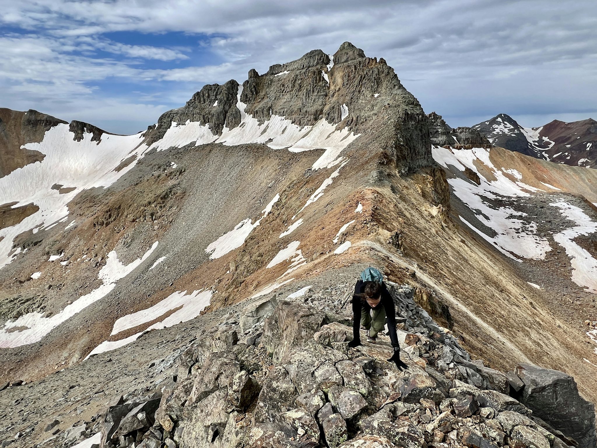 A hiker scrambles up a steep, rocky ridgeline toward the dark jagged summit block of Golden Horn, with snowfields streaking the slopes behind.