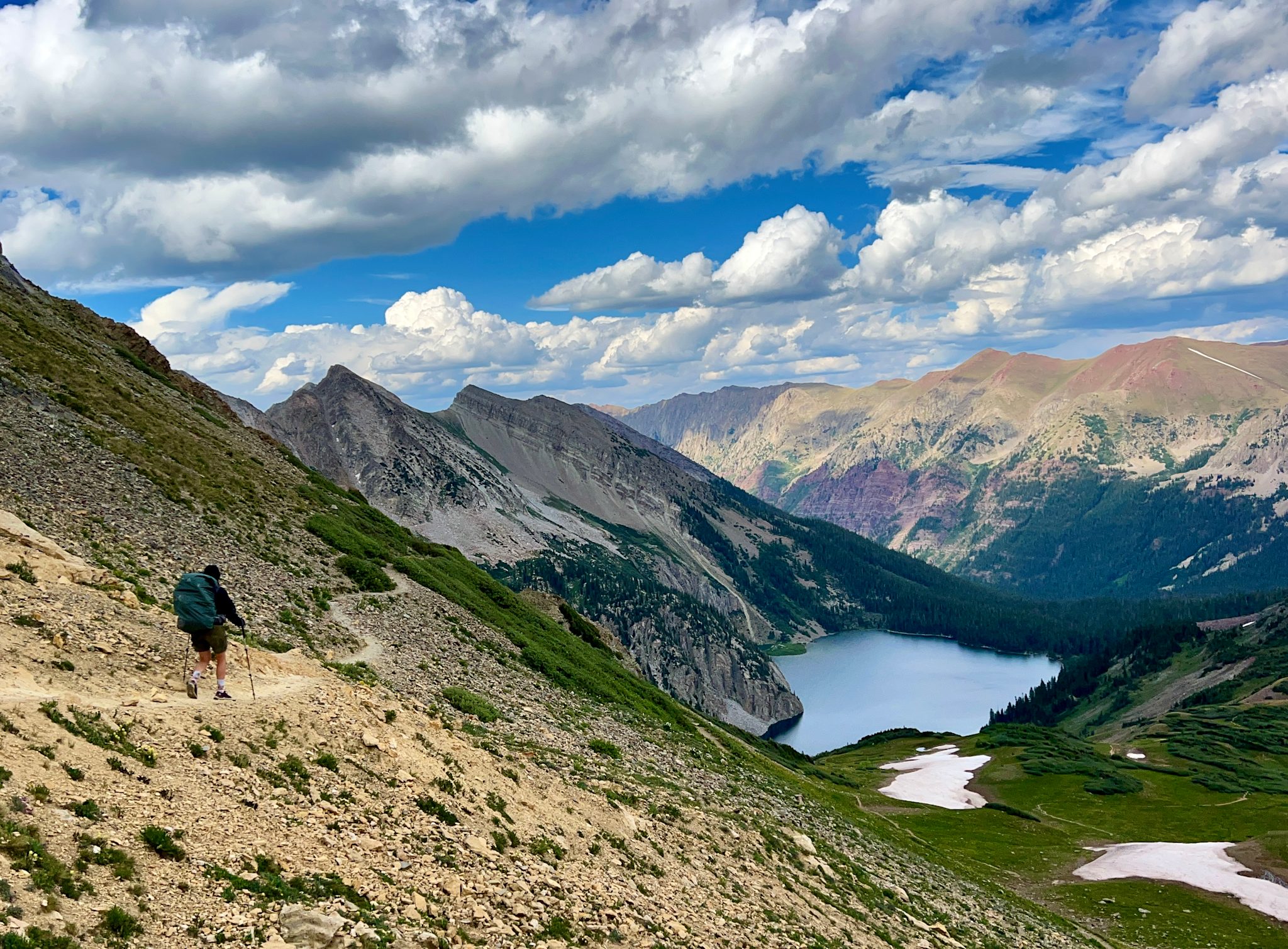 A backpacker with a large pack descends a trail cut across a steep alpine slope, with a turquoise lake in the valley and layered peaks under towering cumulus clouds.