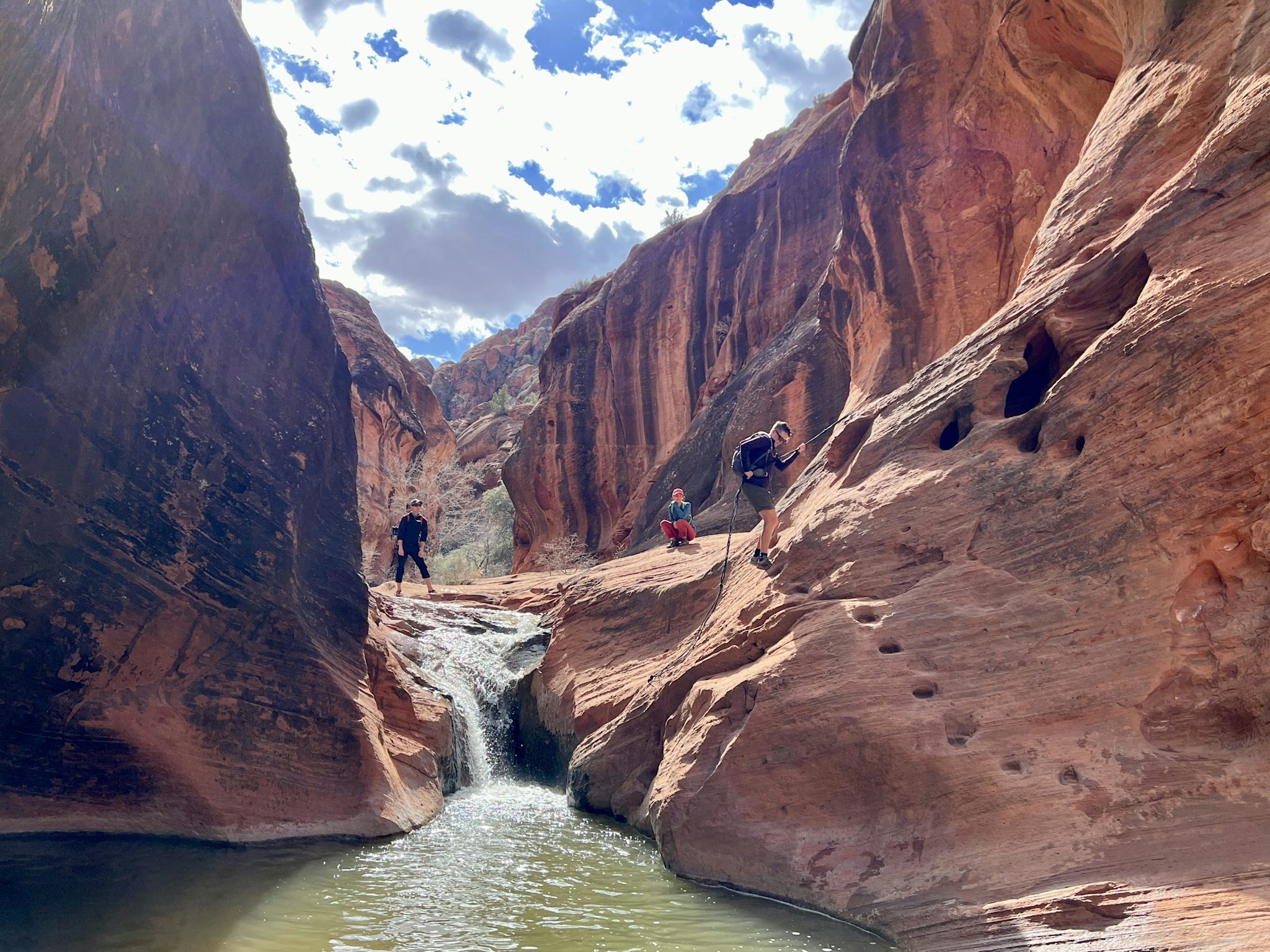 Three hikers stand on ledges above a small waterfall pouring into a green pool, framed by the curving red sandstone walls of a slot canyon.