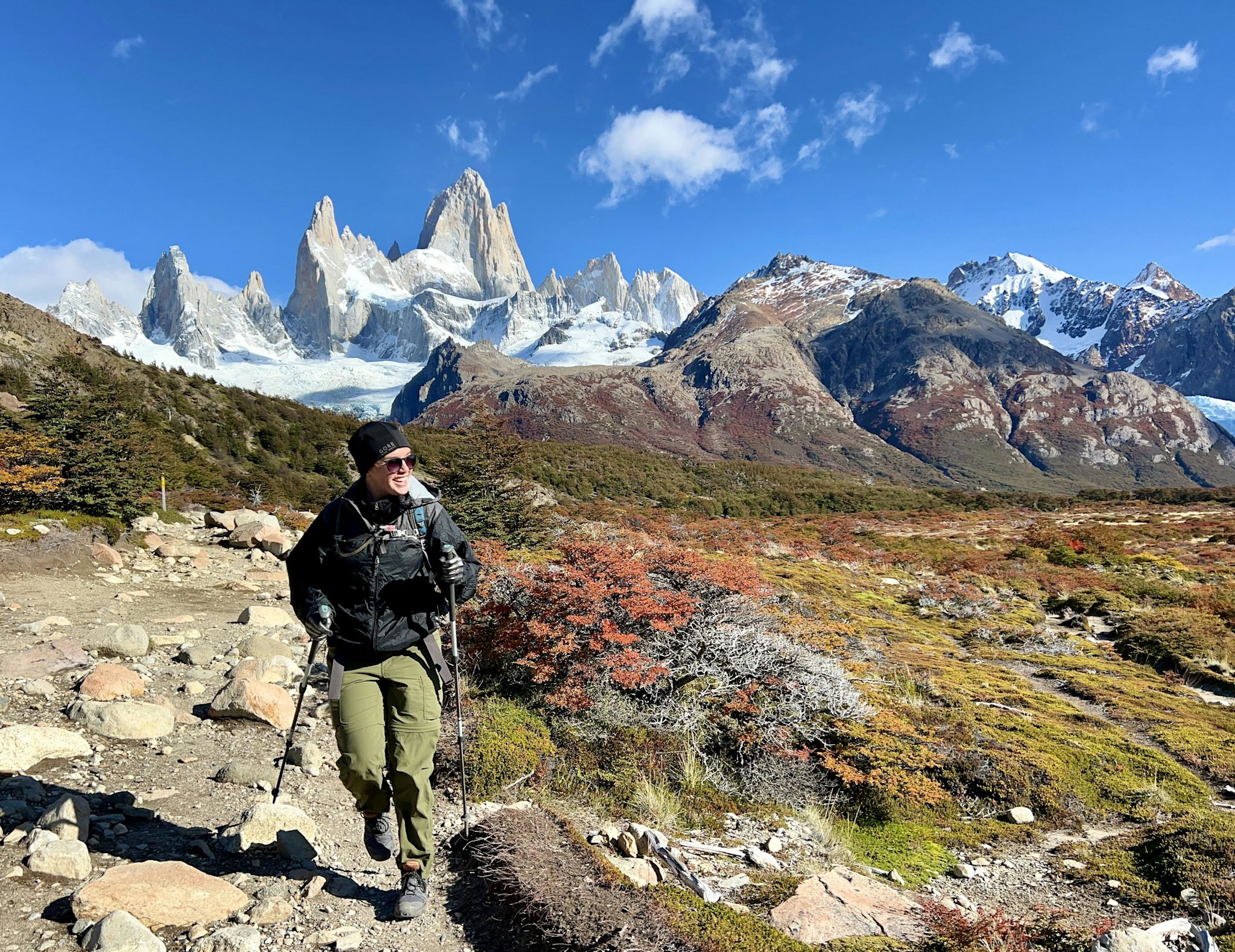 A hiker with trekking poles walks a rocky trail through autumn-colored scrub, with the snow-draped spires of Monte Fitz Roy filling the skyline behind.