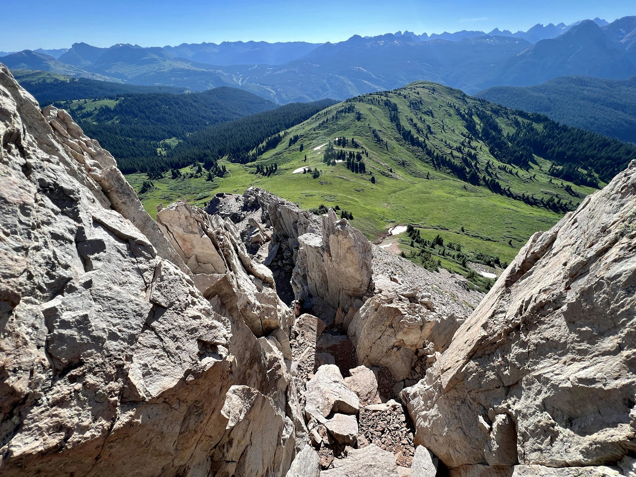 The view down a steep, blocky rock gully from high on a summit ridge, opening onto green alpine meadows and row upon row of distant peaks.