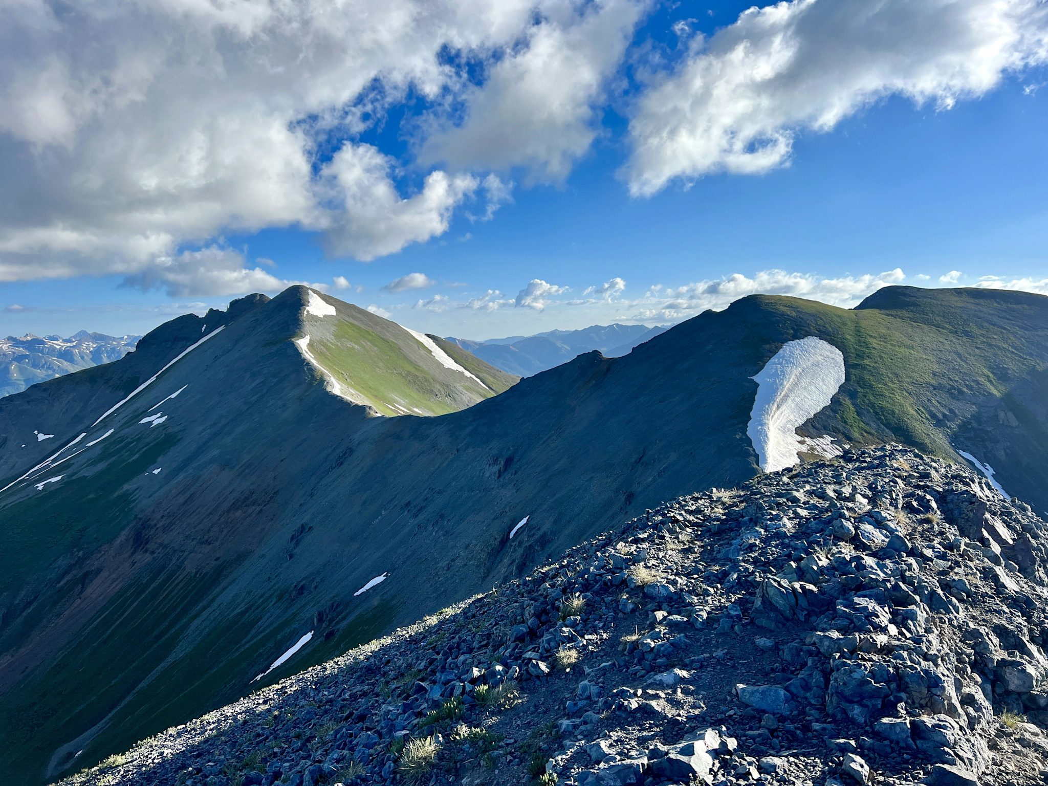 A broad, grassy summit ridge with lingering snow patches curves toward a rounded peak, with the San Juan range receding into the distance under scattered clouds.