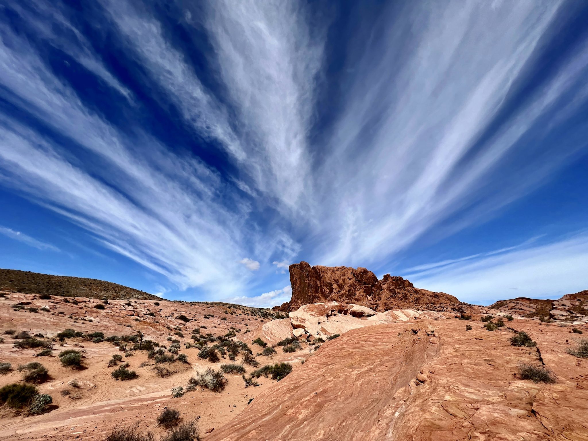 Dramatic wispy cirrus clouds fan across a deep blue sky above the red and cream sandstone formations and desert scrub of Valley of Fire.